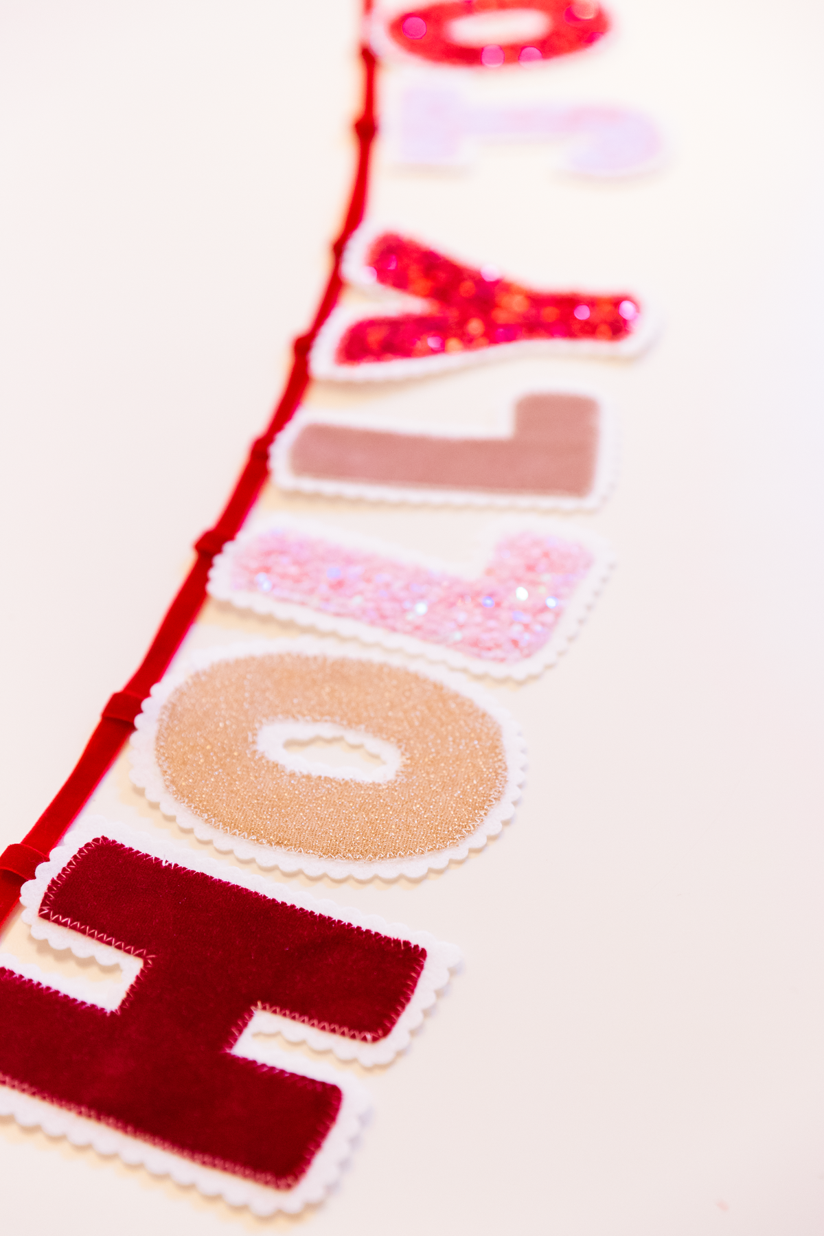 Handmade HOLLY JOLLY Christmas fabric banner featuring pink and red velvet and sequin letters with white scalloped edges, hanging from a red ribbon.